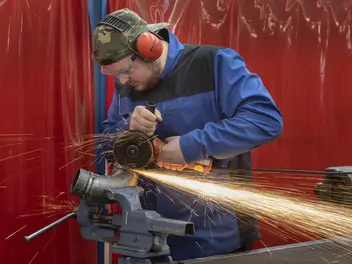 Junger Mann im Blaumann und Ohrschützern flext an einem Rohr, Funken sprühen