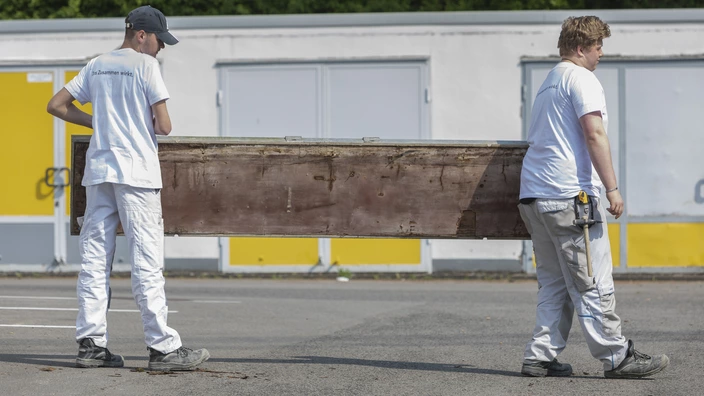Zwei junge Auszubildende in Arbeitskleidung tragen ein großes, rechteckiges Holzbrett. Sie laufen auf einer gepflasterten Fläche, im Hintergrund sind Lagereinheiten zu sehen.