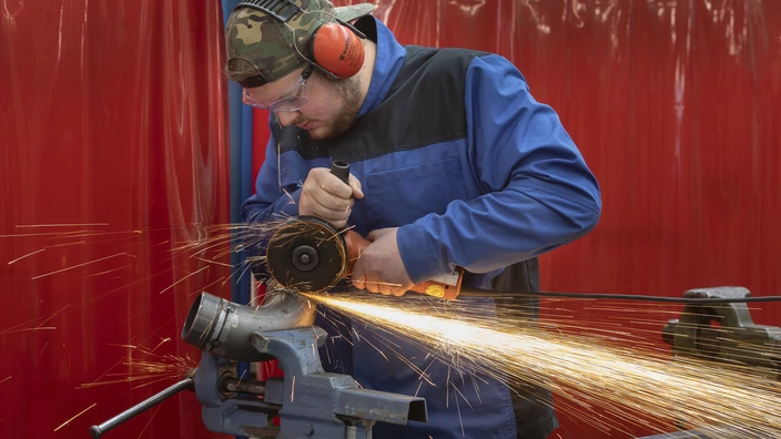 Junger Mann im Blaumann und Ohrschützern flext an einem Rohr, Funken sprühen
